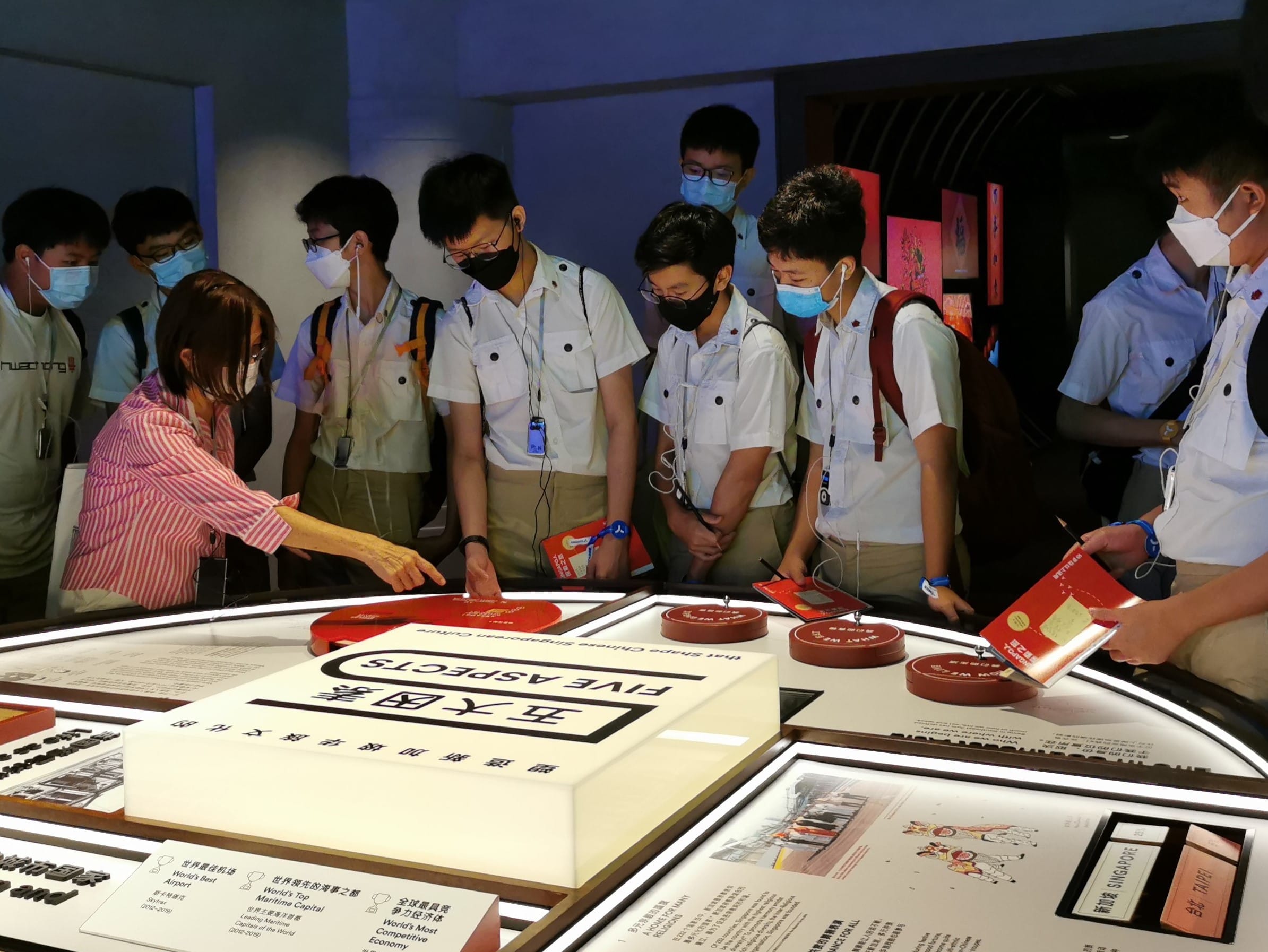 Students in uniforms and masks gather around a lit museum display titled "Five Aspects that Shape Singaporean Culture."